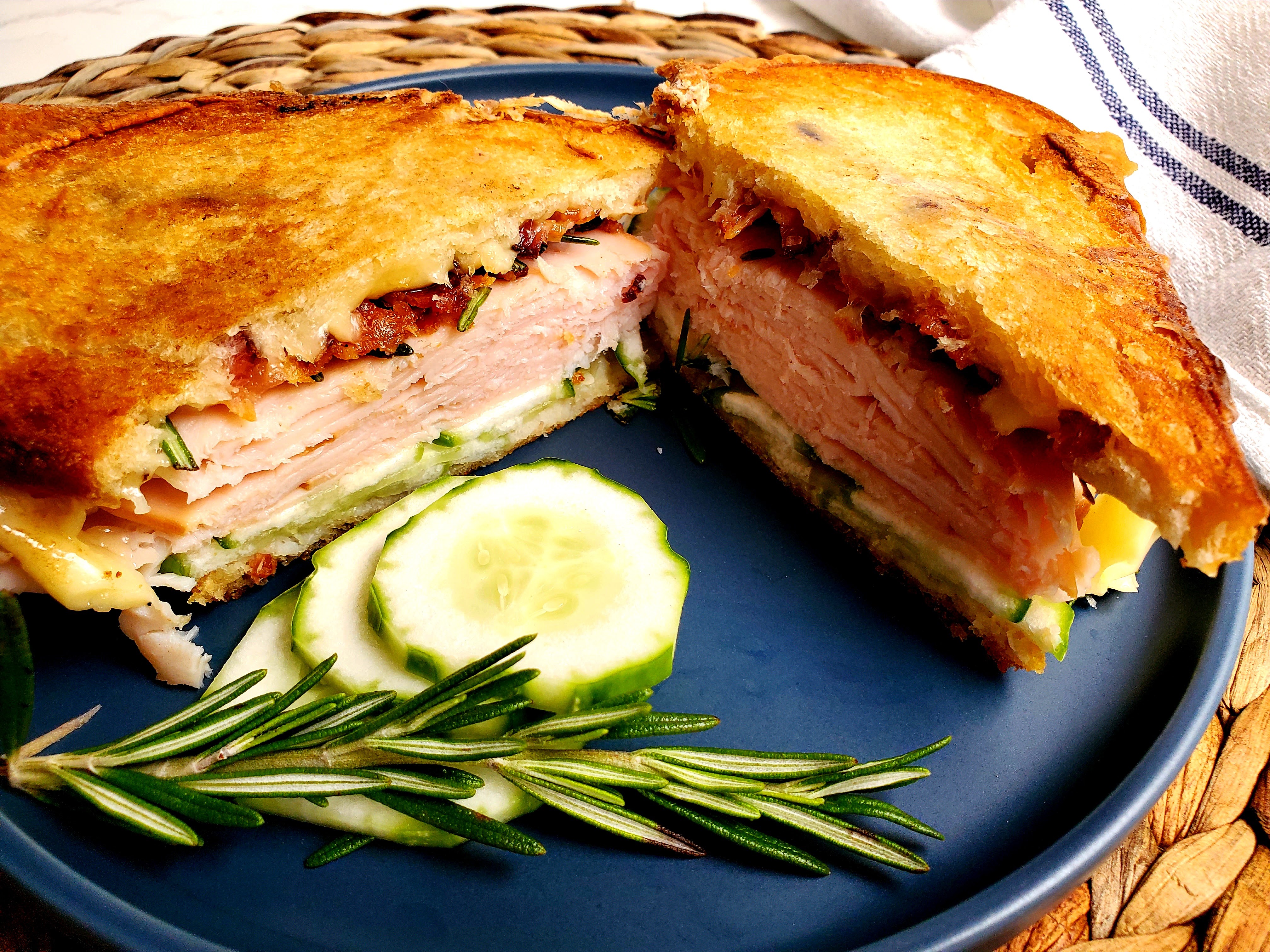 sanwich with turkey, bacon, cucumber, fresh rosemary and cheese sit on a plate with a sprig pf rosemary and sliced cucumber