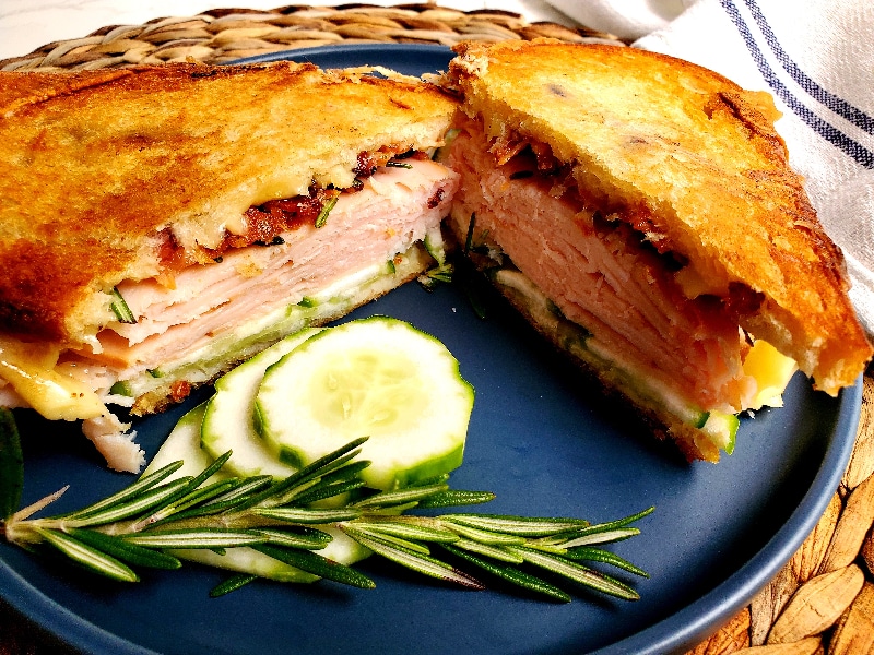 a sandwich made of turkey, bacon, cucumber, cheese, and rosemary sit on a plate with a sprig opf rosemary and sliced cucumber